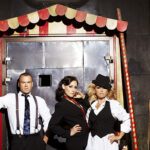 Band Alcazar steht vor einem Jahrmarktstand, ein Mann mit Hosenträgern und Krawatte daneben zwei Frauen ebenfalls in Anzügen, eine trägt einen Hut