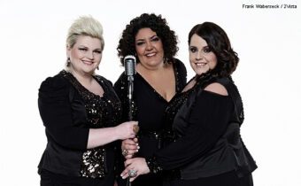 Band BigSoul in schwarzer glitzernder Kleidung, drei Frauen mit kurzen Haaren stehen vor einem Mikrofon