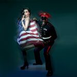 Sänger Captain Jack in einem schwarzen Offiziersanzug mit roter Mütze und einer Sonnenbrille, hält eine amerikanische Fahne die eine Frau als Kleid trägt