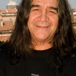 Sänger Drupi steht auf einem Balkon, eine Stadt im Hintergrund, er trägt ein schwarzes Shirt und hat lange Haare