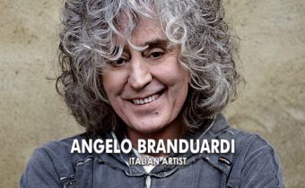 Sänger Angelo Branduardi steht vor einem braunen Hintergrund, hat graue Locken und einen grauen Pullover an