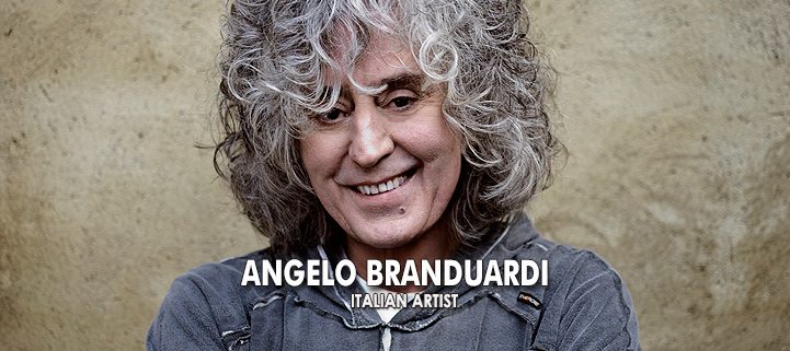 Sänger Angelo Branduardi steht vor einem braunen Hintergrund, hat graue Locken und einen grauen Pullover an