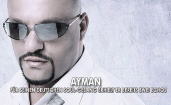 Sänger Ayman blickt nach unten, trägt ein weißes Hemd mit Krawatte, er trägt eine Sonnenbrille und steht vor grauem Hintergrund