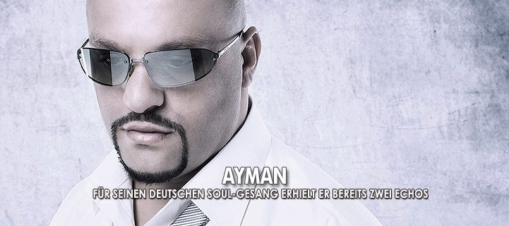 Sänger Ayman blickt nach unten, trägt ein weißes Hemd mit Krawatte, er trägt eine Sonnenbrille und steht vor grauem Hintergrund