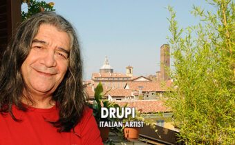 Sänger Drupi steht auf einem Balkon, eine Stadt im Hintergrund und ein Baum, er trägt ein rotes Shirt und hat lange Haare