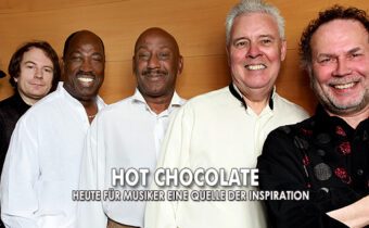 Band Hot Chocolate steht vor einer Holzwand, die Männer lächeln