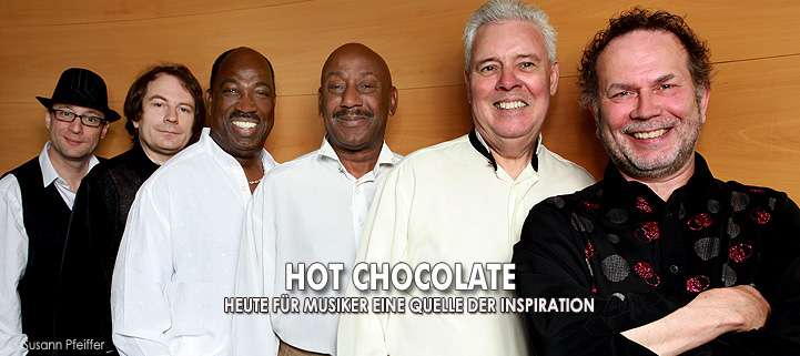 Band Hot Chocolate steht vor einer Holzwand, die Männer lächeln