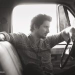 Sänger TomBeck in einem Auto mit Blick aus der Windschutzscheibe und Hand am Lenkrad in schwarz weiß
